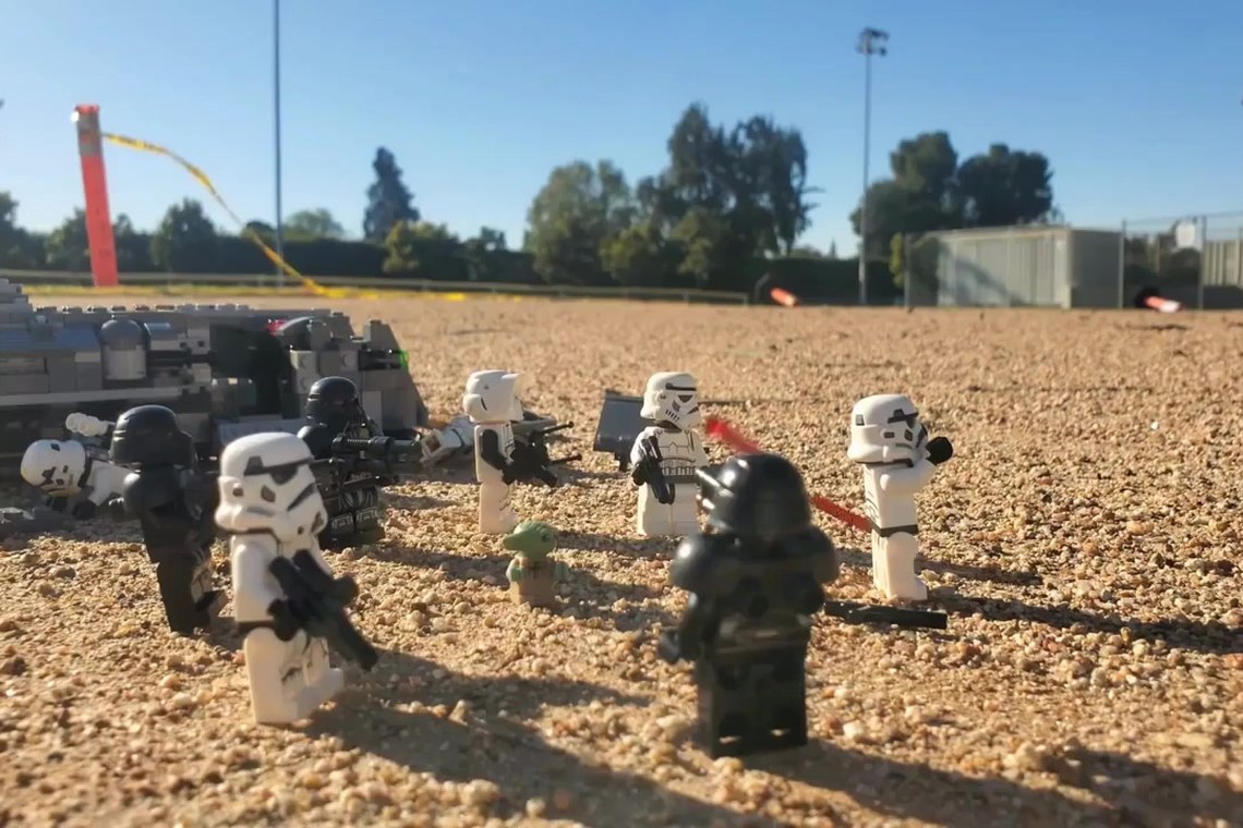 Lego stormtroopers advancing across gravel on a school field, a blaster bolt in flight