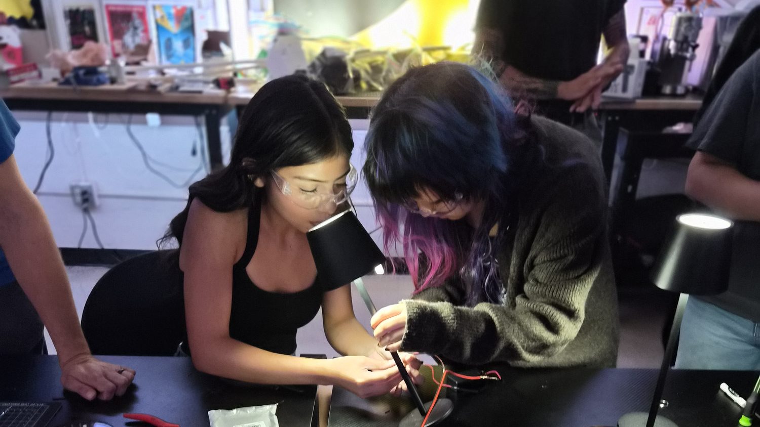 Two students wiring and heat-shrinking a circuit at the bench under a work lamp